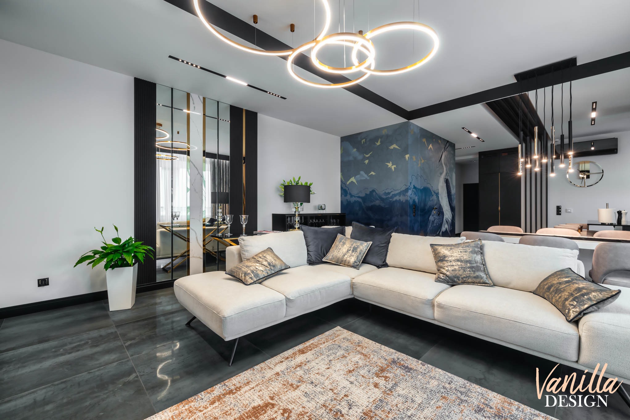 Nowoczesny salon z narożną sofą w odcieniach beżu, dekoracyjnym dywanem, lustrzaną ścianą z czarnymi akcentami i designerskim oświetleniem.