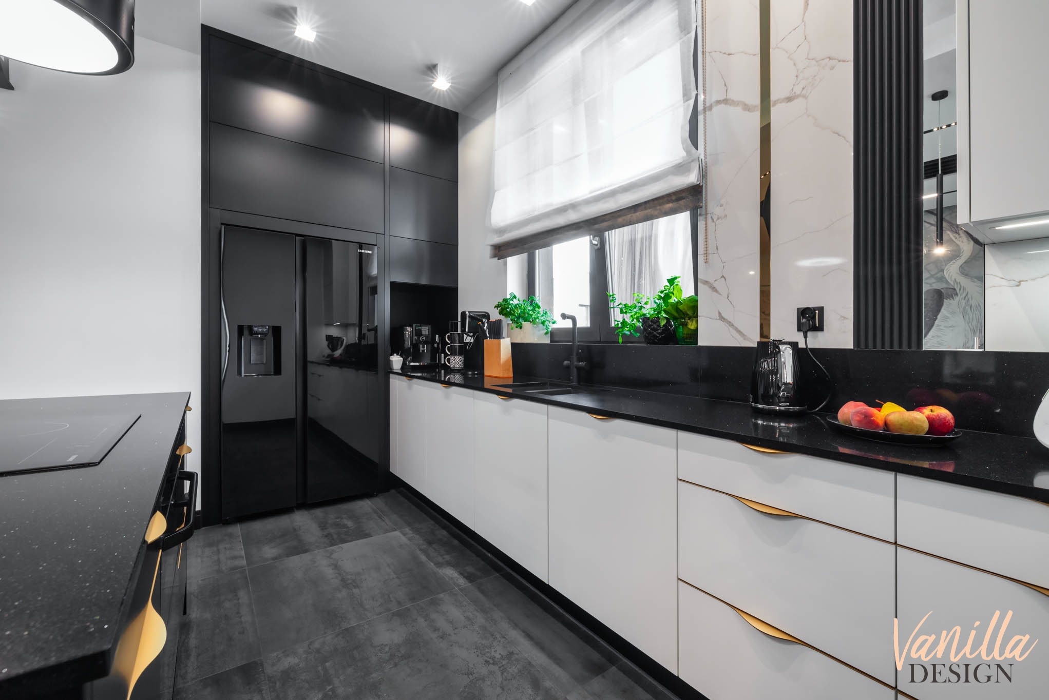 Nowoczesna kuchnia z czarnymi frontami lodówki i szafek górnych kontrastującymi z białymi szafkami dolnymi z złotymi uchwytami, czarnym blatem i szarymi płytkami podłogowymi.