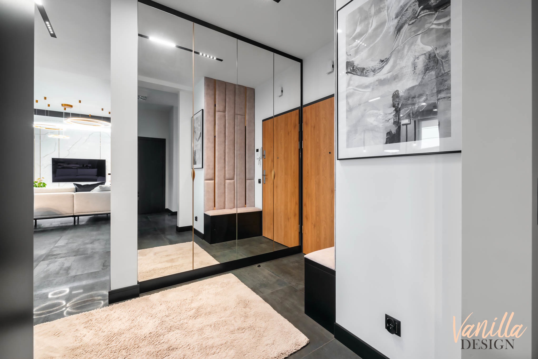 Nowoczesny przedpokój z dużym lustrem, panelami tapicerowanymi, drewnianymi drzwiami i abstrakcyjnym obrazem w czarnej ramie.