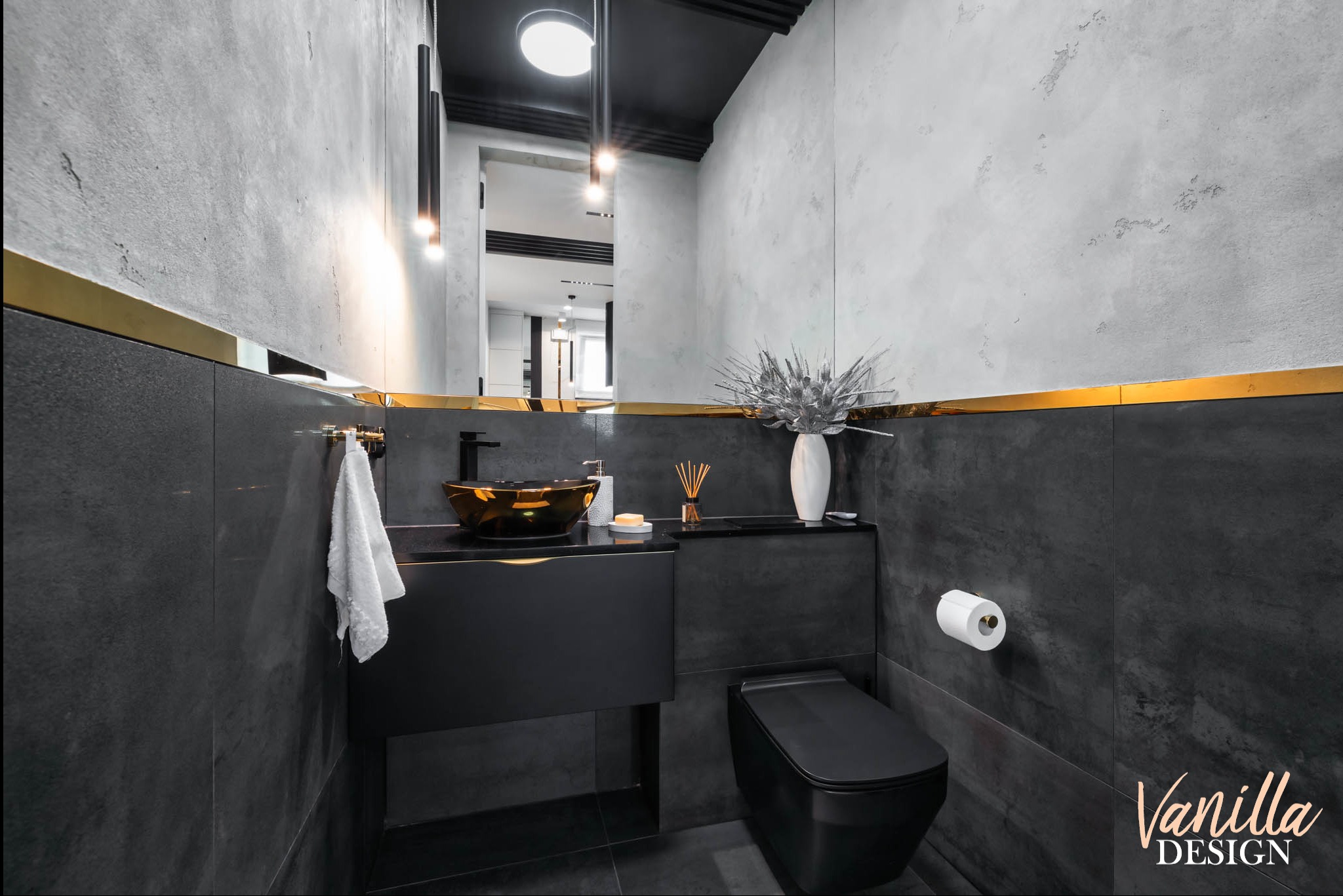 Nowoczesna łazienka w odcieniach szarości i czerni z akcentami złota, umywalka w kolorze starego złota, minimalistyczna toaleta, dekoracyjny wazon z suchymi gałązkami, lustro z oświetleniem.
