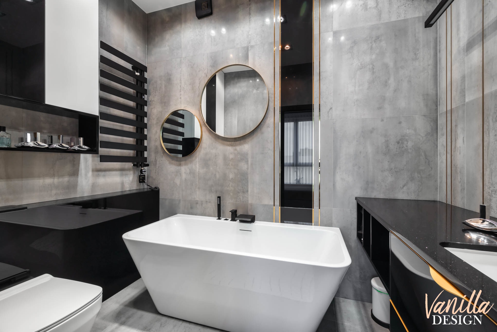 Nowoczesna łazienka z wolnostojącą białą wanną, szarą płytką imitującą beton, okrągłymi lustrami w złotych ramach i czarnymi akcentami, takimi jak grzejnik drabinkowy i blat umywalkowy.