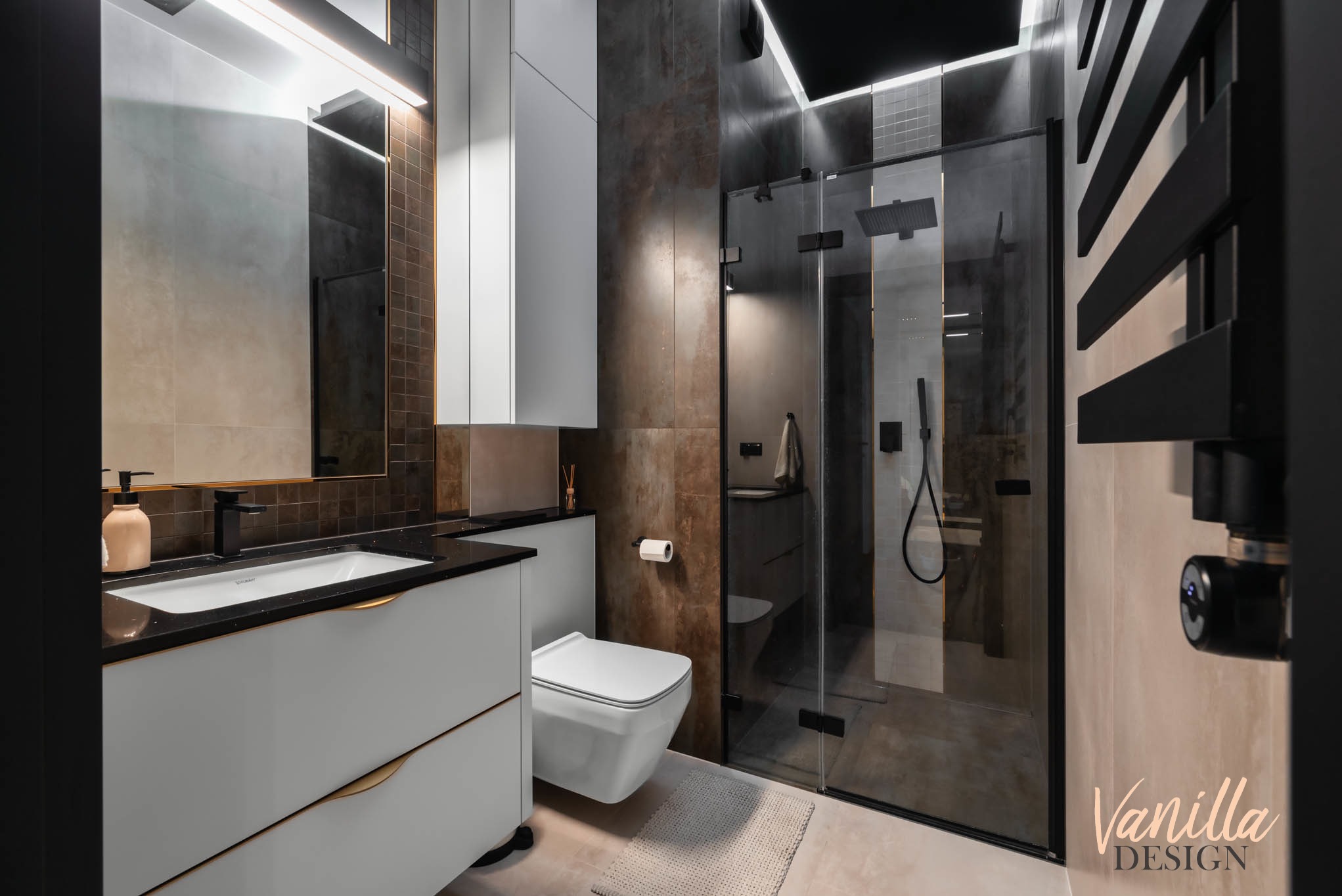 Nowoczesna łazienka w odcieniach brązu i szarości, z kabiną prysznicową typu walk-in, podwieszaną toaletą i umywalką na szafce z czarnym blatem, oświetlona listwą LED nad lustrem.