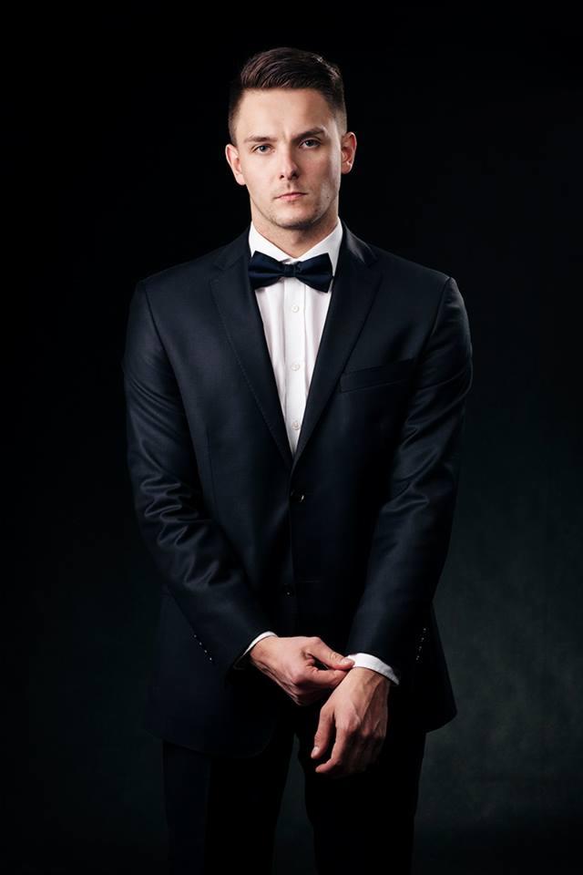 Mężczyzna w czarnym garniturze i muszce na ciemnym tle, z rękami złożonymi przed sobą, patrzący wprost w obiektyw.