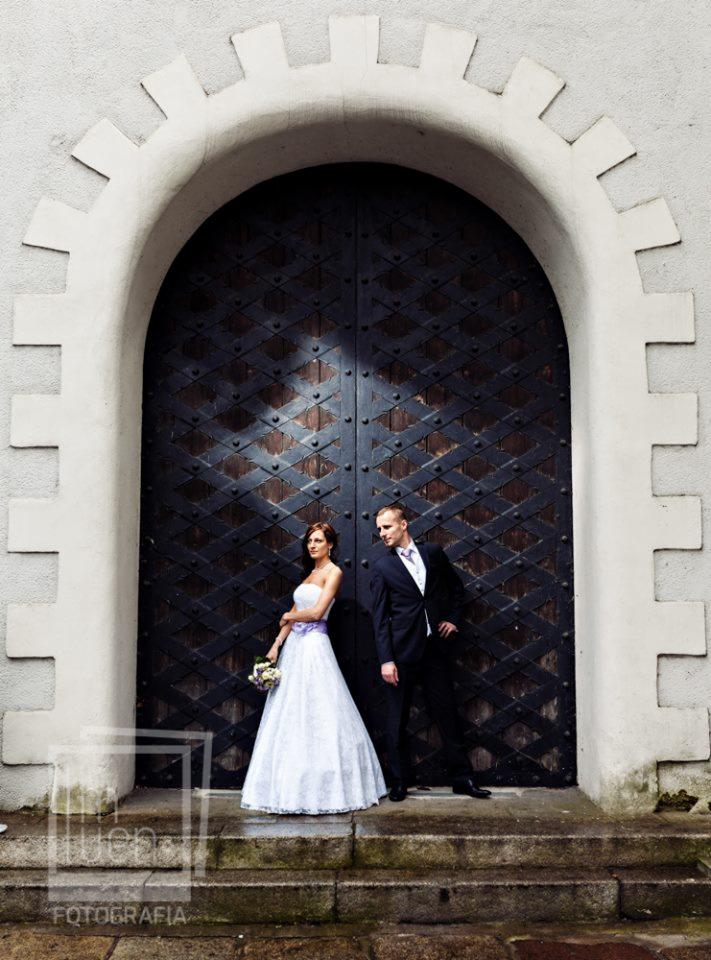 Para młoda pozuje na tle ogromnych, ciemnych drzwi z metalowymi zdobieniami i kamiennego łuku wejściowego. Panna młoda w białej sukni z fioletową szarfą trzyma bukiet. Pan młody w ciemnym...