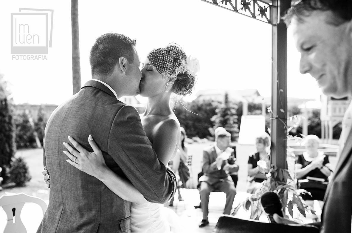 Czarno-białe zdjęcie pary młodej całującej się pod ozdobnym baldachimem, widoczni goście w tle, panna młoda w siateczkowym welonie i z kwiatem we włosach, pierścionek na palcu.