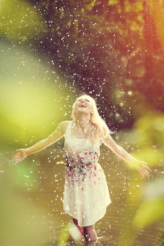 Uśmiechnięta blondynka w kwiecistej sukience stoi w płytkiej wodzie, rozpościerając ramiona w geście radości, otoczona kropelkami wody w promieniach słońca.