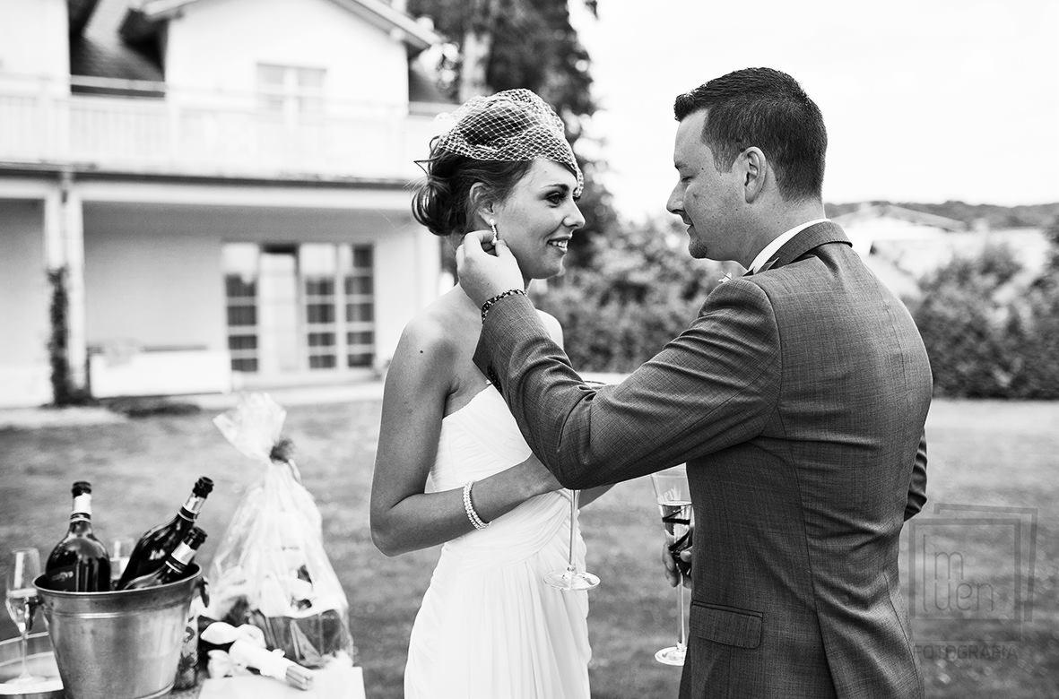 Czarno-białe zdjęcie pary młodej podczas ceremonii: panna młoda w sukni bez ramiączek i siatkowym welonie, pan młody w garniturze poprawia jej kolczyk, w tle dom i wiadro z szampanem.