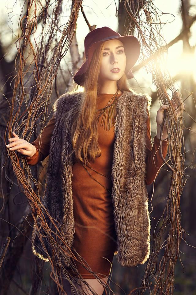 Portret kobiety w brązowej sukience i futrzanej kamizelce, stojącej wśród suchych gałęzi na tle zachodzącego słońca, z kapeluszem na głowie i długim naszyjnikiem.