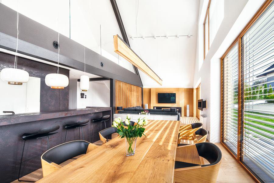Jasne, przestronne wnętrze domu z wysokim sufitem, drewnianym stołem, krzesłami z czarnymi siedziskami, kwiatami w wazonie, barem z hokerami i dużymi oknami z żaluzjami.