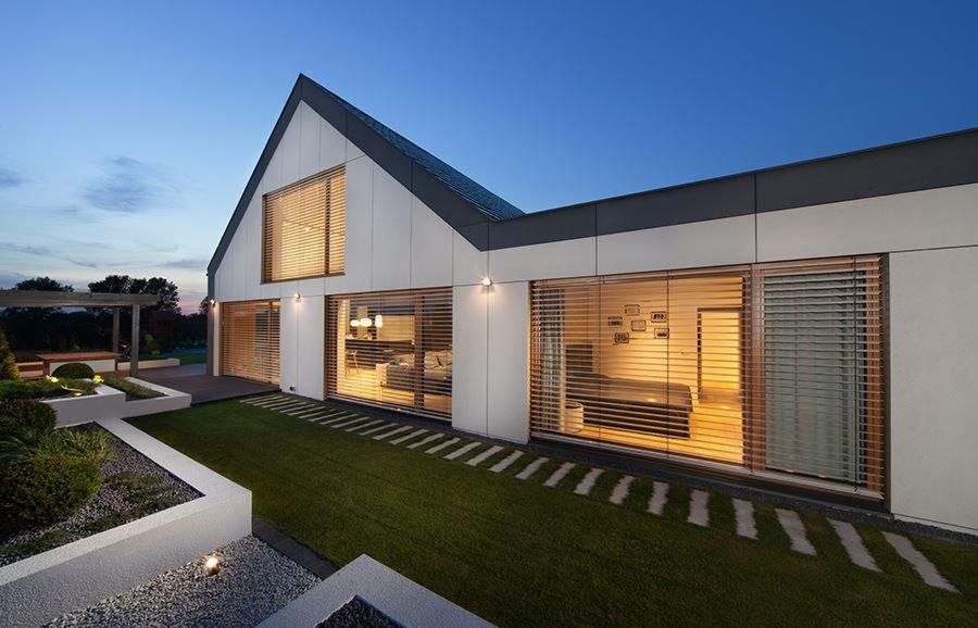 Nowoczesny dom jednorodzinny z ogrodem o zmierzchu, widok z zewnątrz przez duże okna z żaluzjami na wnętrze salonu i sypialni, ścieżka z płyt na trawniku.