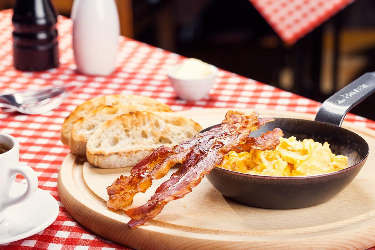 Śniadanie na drewnianej desce: jajecznica w patelni, chrupiący bekon, tosty, kawa i masło na tle czerwono-białej kraty.