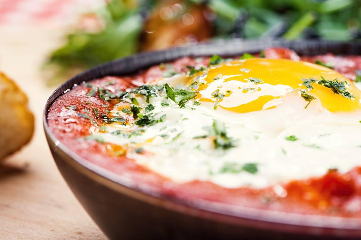 Jajko sadzone w pomidorach, posypane świeżą zieloną pietruszką, podane w czarnej patelni na drewnianym stole, z boku kromka chleba.