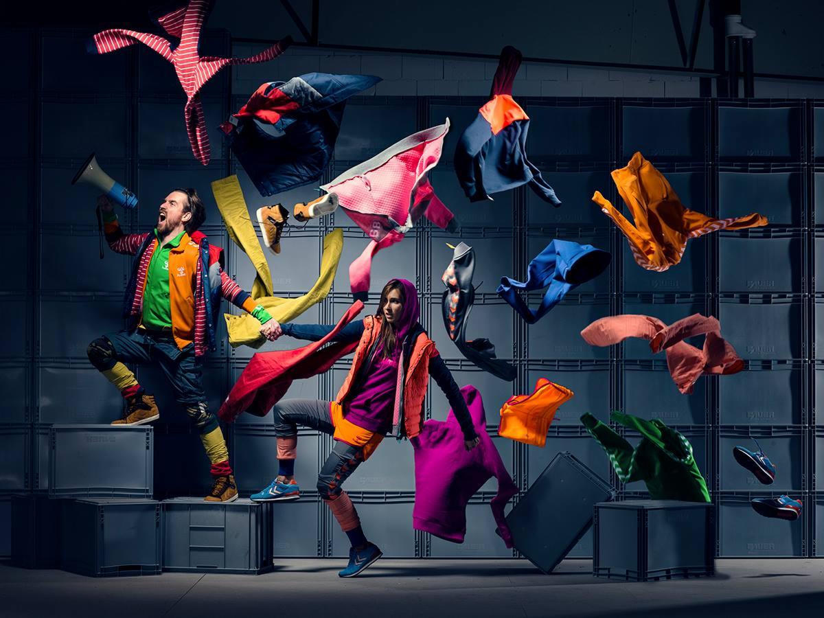 Dynamiczne ujęcie dwojga modeli w ruchu, otoczonych unoszącymi się ubraniami i butami na tle ściany z szarych skrzyń; mężczyzna krzyczy przez megafon, kobieta w pomarańczowej kurtce pozuje w locie.