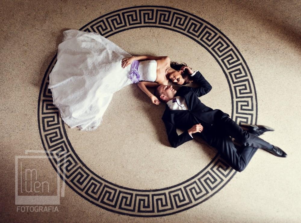 Nowożeńcy w Szczecinie leżący na podłodze z geometrycznym wzorem, panna młoda w białej sukni z fioletową szarfą, pan młody w czarnym garniturze, ujęcie z góry.