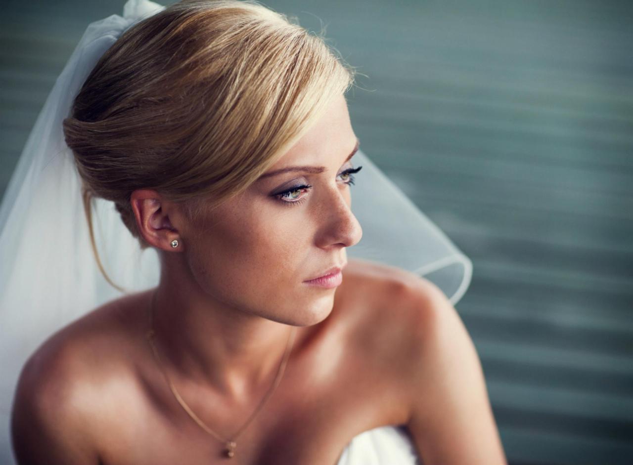 Profil panny młodej z blond włosami upiętymi w kok, delikatny makijaż, welon i złoty łańcuszek, spoglądającej w lewo.