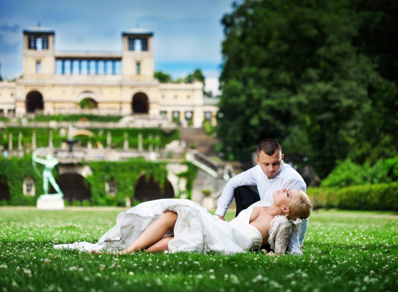 Panna młoda w białej sukni leży na trawie, a pan młody w białej koszuli klęczy obok niej, z budynkiem w tle.