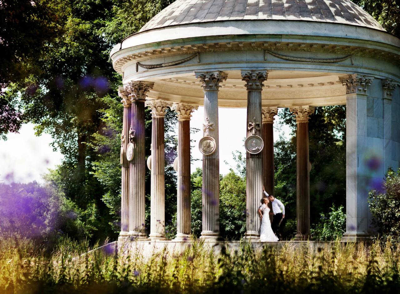 Para młoda w objęciach pod klasycystyczną altaną z kolumnami ozdobionymi medalionami, otoczoną zielenią parku.