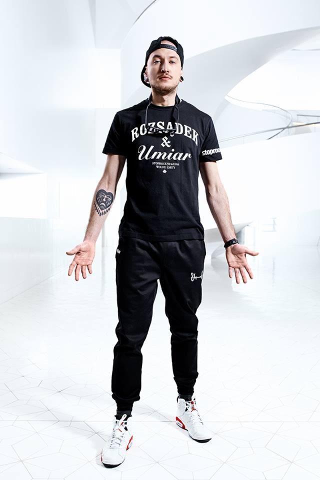 Mężczyzna w czarnym stroju z tatuażem na ramieniu, stojący na białej, geometrycznej podłodze, z rozłożonymi rękami, w jasnym, minimalistycznym wnętrzu.