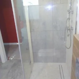 Kabina prysznicowa z odpływem liniowym w nowocześnie wykończonej łazience, widoczne szare płytki imitujące kamień na ścianach i mozaika na podłodze, zarys czerwonej ściany w tle.