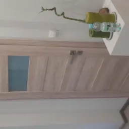 Drewniane drzwi wewnętrzne z prostokątnym przeszkleniem, widoczne z boku, z dekoracjami na szafce obok.