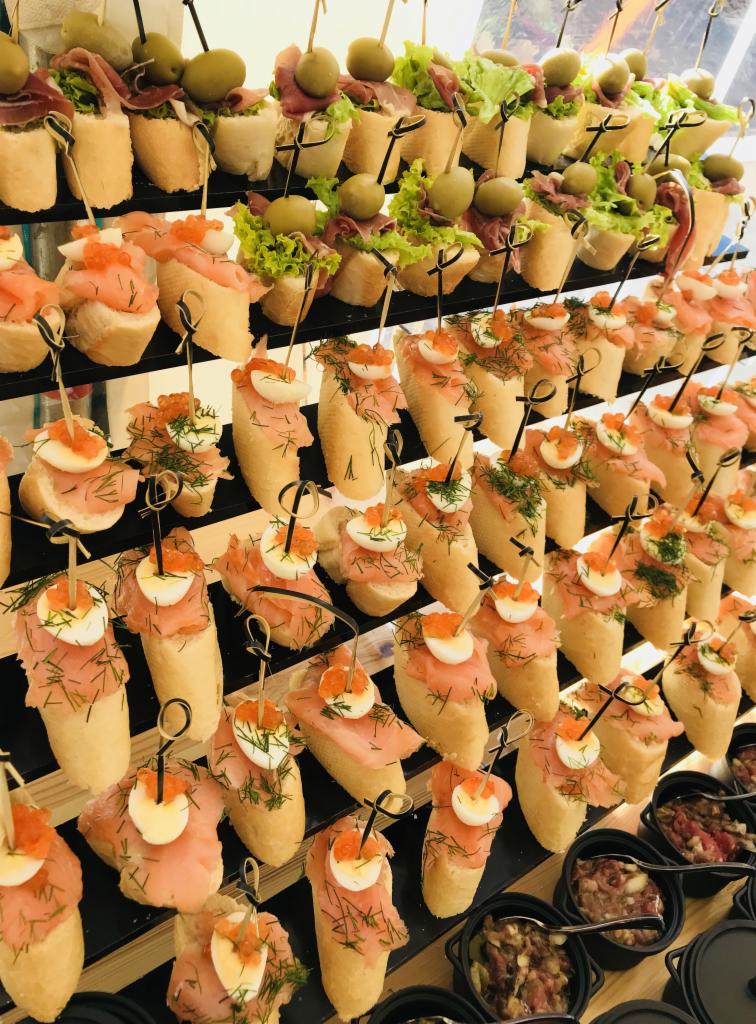 Elegancko ułożone kanapeczki na czarnym stojaku: górny rząd z szynką parmeńską, sałatą i oliwką, poniżej kanapki z łososiem, jajkiem przepiórczym i kawiorem.