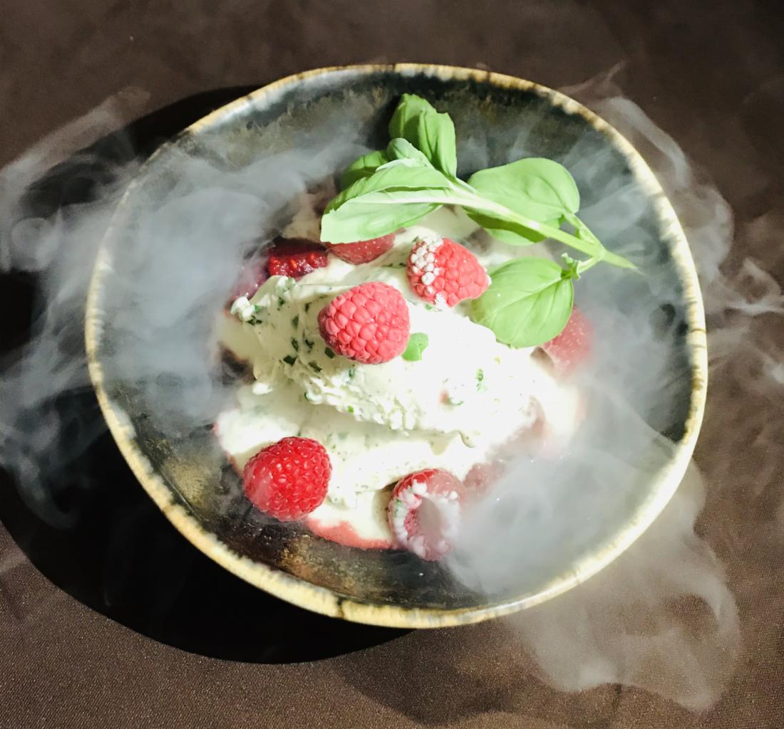 Deser lodowy z malinami i bazylią w ceramicznej misce otoczony dymem, na ciemnym tle.