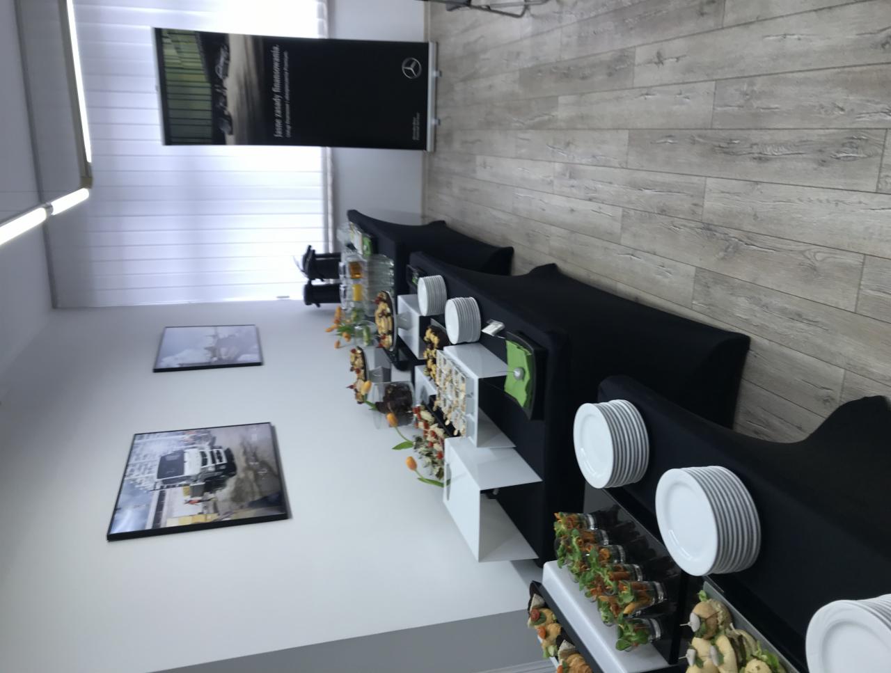 Długi stół cateringowy przy ścianie z dekoracjami, zastawiony przekąskami i stosem białych talerzy, z banerem reklamowym w tle.