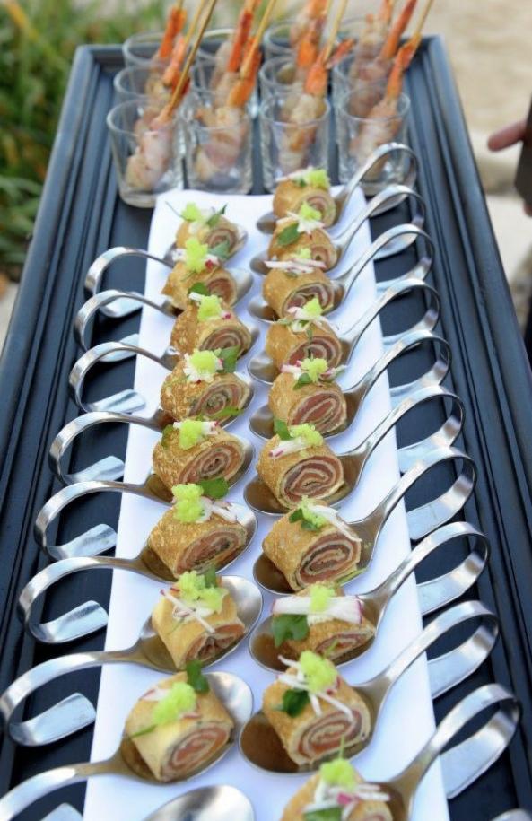 Ekskluzywny catering: Rząd srebrnych łyżek z roladkami z łososia i dekoracją z zielonego kawioru, obok szklanek z krewetkami na szpadce, ułożonych na czarnej tacy.