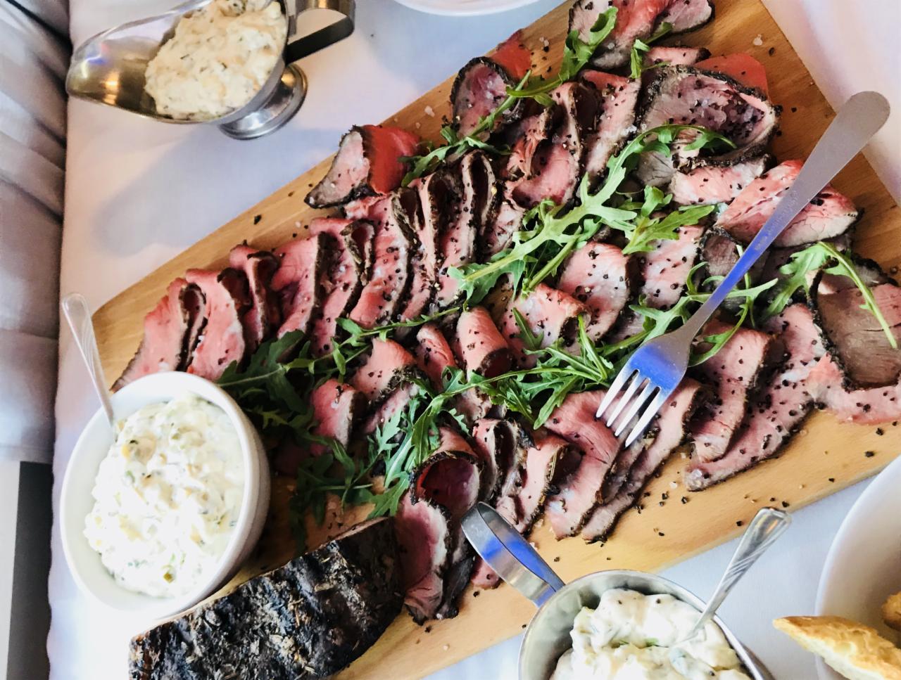 Deska z plastrami pieczonej wołowiny posypanej pieprzem, udekorowana rukolą, serwowana z trzema sosami na białym obrusie, widoczny widelec do nakładania.