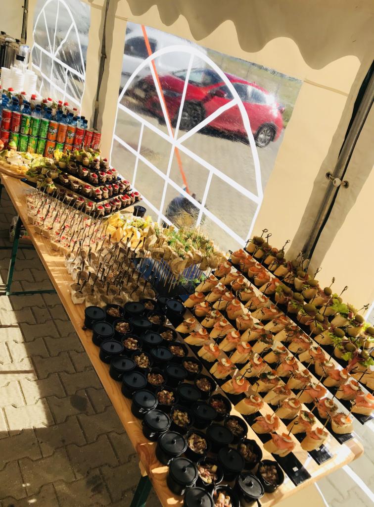 Stół cateringowy w Gdańsku pod namiotem, bogato zastawiony różnorodnymi przekąskami: mini-dania w czarnych naczyniach, koreczki, kanapeczki z łososiem, napoje w tle. Widoczny fragment czerwonego...