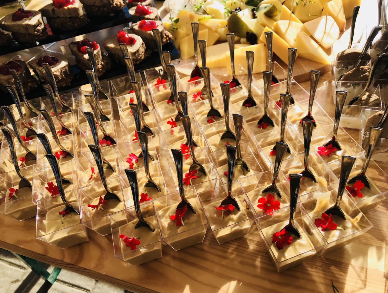 Ustawione rzędem porcje kremowego deseru w przezroczystych naczyniach z łyżeczkami i czerwonymi kwiatami, na drewnianym blacie, w tle sery i przekąski z pasztetem i żurawiną.