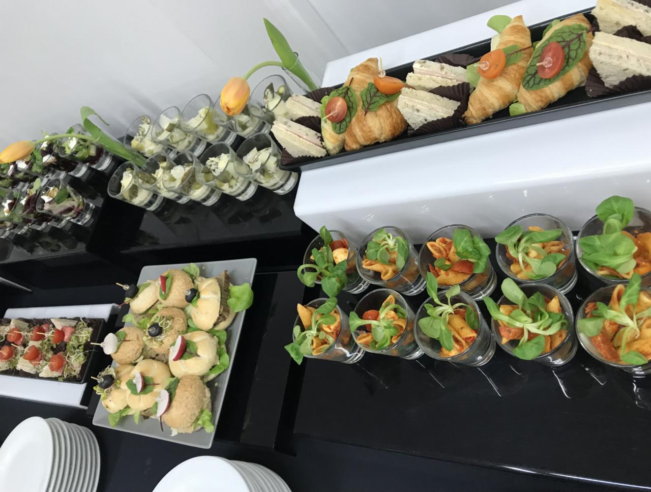 Elegancko ułożone przekąski cateringowe na czarnym bufecie: mini kanapki, croissanty z pomidorem i bazylią, sałatki w szklankach, dekorowane tulipanami.