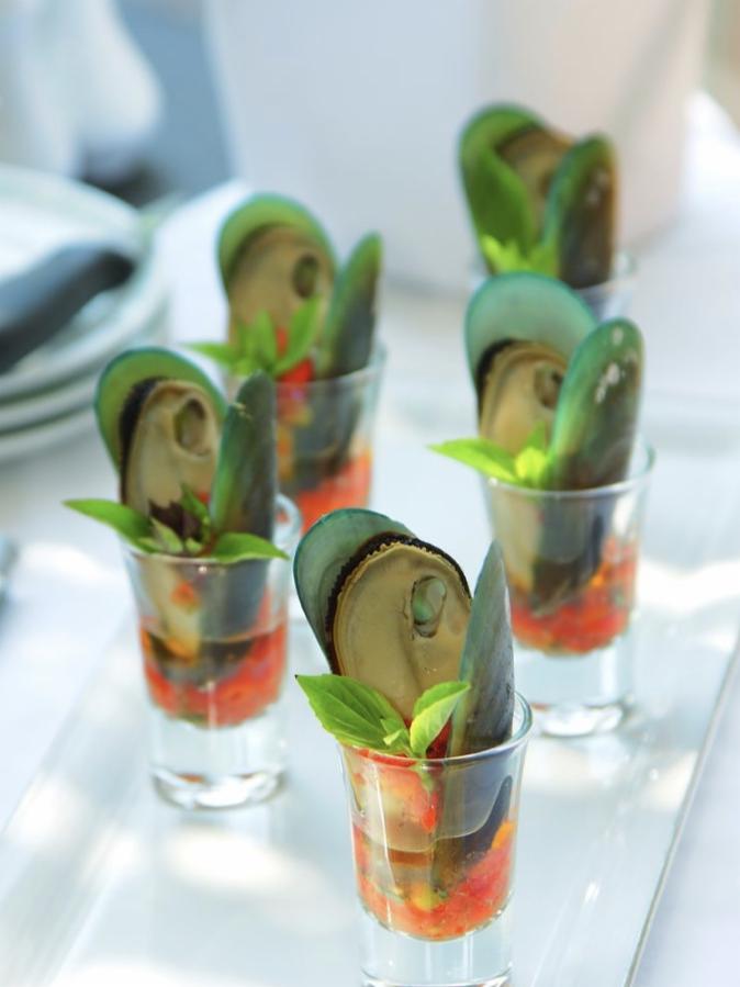 Eleganckie przekąski: Szklanki z małżami nowozelandzkimi, sosem pomidorowym i listkami bazylii, ułożone na białym stole.