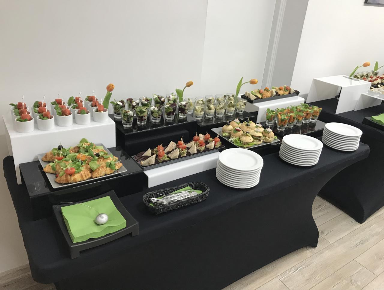 Elegancki stół cateringowy z różnorodnymi przekąskami: mini sałatki w szklankach, croissanty z warzywami, kanapki, koreczki, talerze i serwetki, całość udekorowana tulipanami.