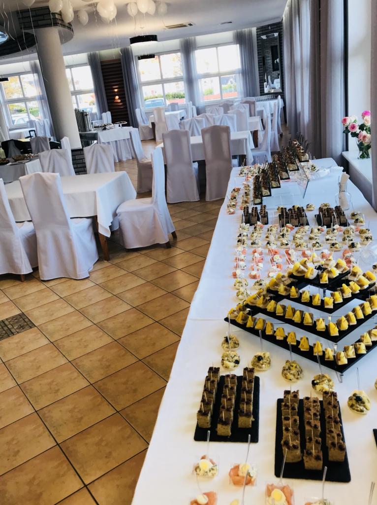 Sala bankietowa przygotowana na przyjęcie, z białymi krzesłami i stołami, oraz stołem szwedzkim pełnym różnorodnych przekąsek, w tym piramidy serowe i kanapeczki na ciemnym pieczywie.