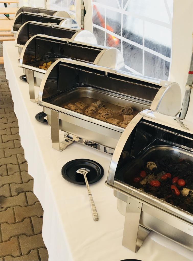 Bufet cateringowy z podgrzewaczami, zawierający różnorodne dania: pieczone ziemniaki, mięso w sosie i warzywa z pomidorami, z czarnym talerzem i chochlą na białym obrusie.