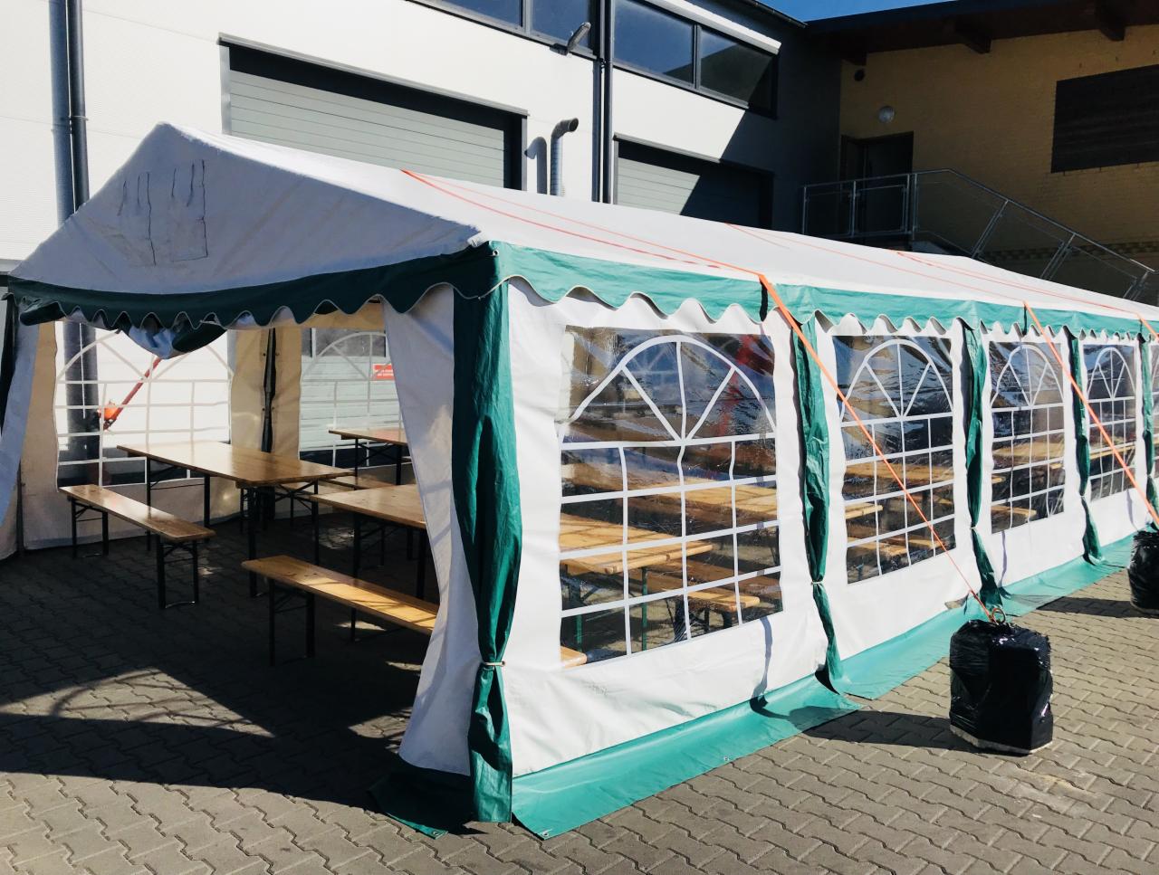 Rozłożony biało-zielony namiot cateringowy z przezroczystymi oknami i drewnianymi stołami z ławkami w środku, zabezpieczony obciążnikami na zewnątrz, na tle budynku.