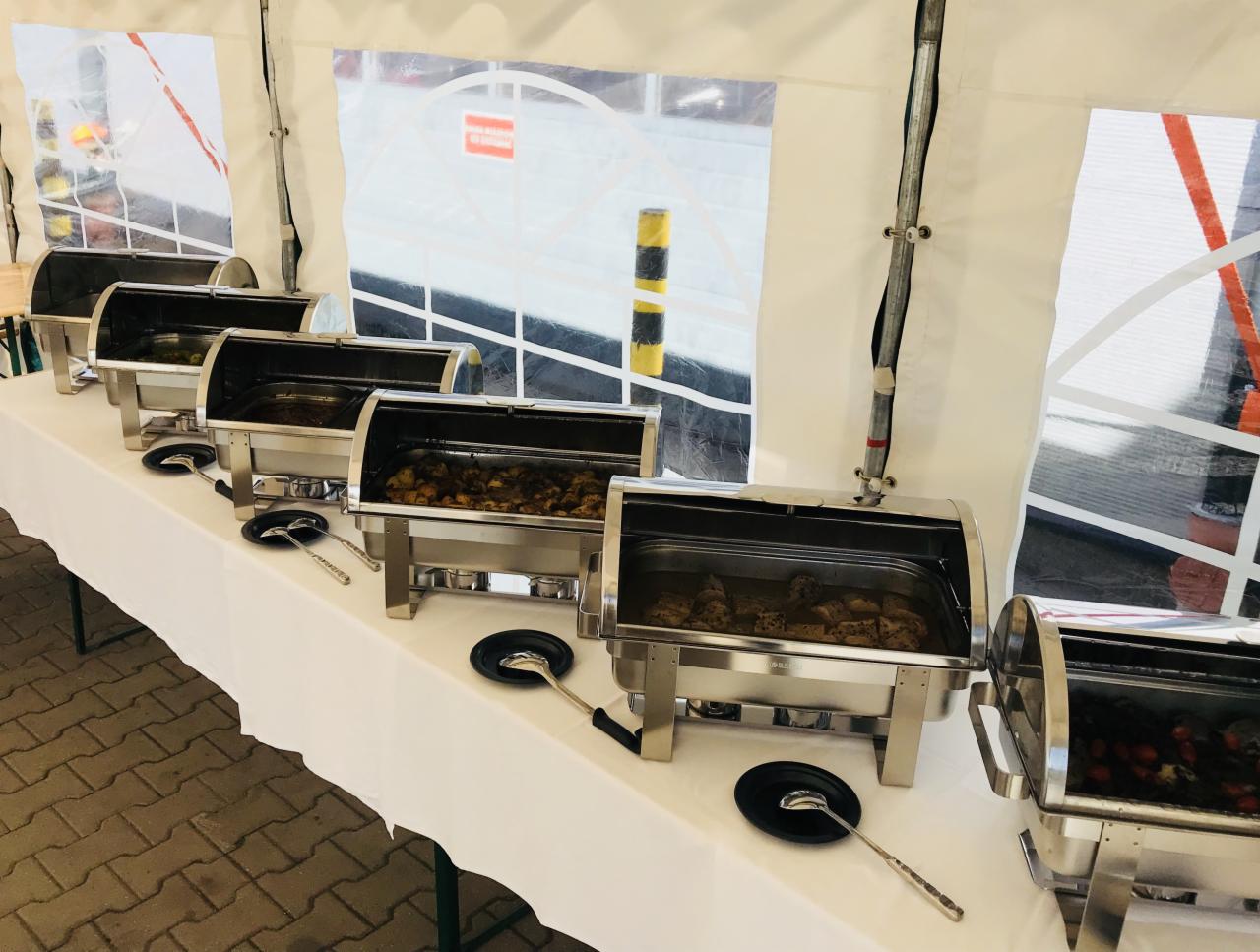 Bufet cateringowy z podgrzewaczami i potrawami mięsnymi oraz warzywnymi pod namiotem, z widoczną konstrukcją namiotu i otoczeniem na zewnątrz.