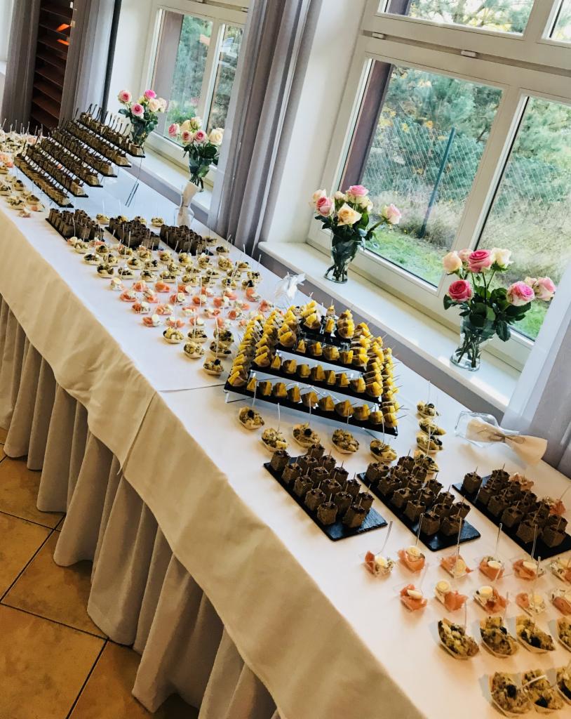 Stół z białym obrusem, na którym znajdują się różnorodne przekąski na paterach i w małych naczyniach, ustawione rzędami; w tle okna z widokiem na zieleń i wazony z różowymi różami.