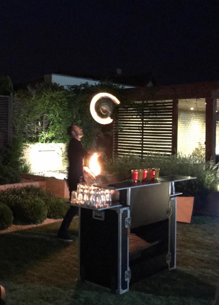 Mężczyzna w czarnym stroju żongluje płonącymi pochodniami na tle baru z kieliszkami i drinkami podczas wieczornego pokazu w ogrodzie.