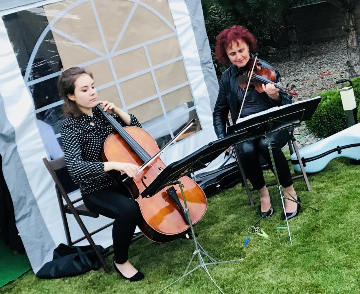 Dwie kobiety grające na instrumentach smyczkowych (wiolonczela i skrzypce) na tle białego namiotu podczas wydarzenia plenerowego.