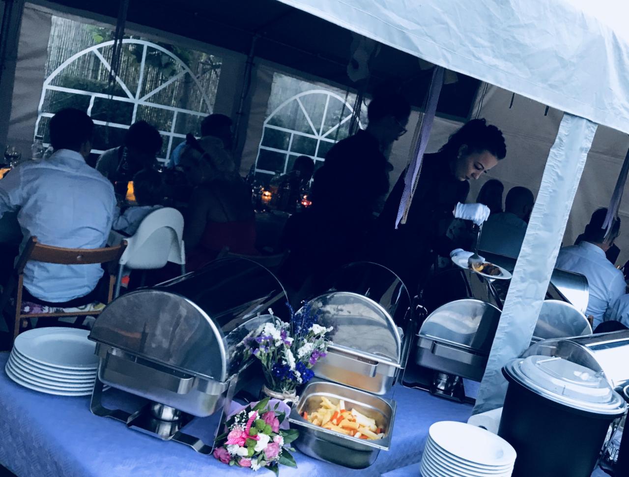 Bufet z podgrzewaczami potraw, dekoracjami kwiatowymi i obsługą w tle podczas przyjęcia pod namiotem.