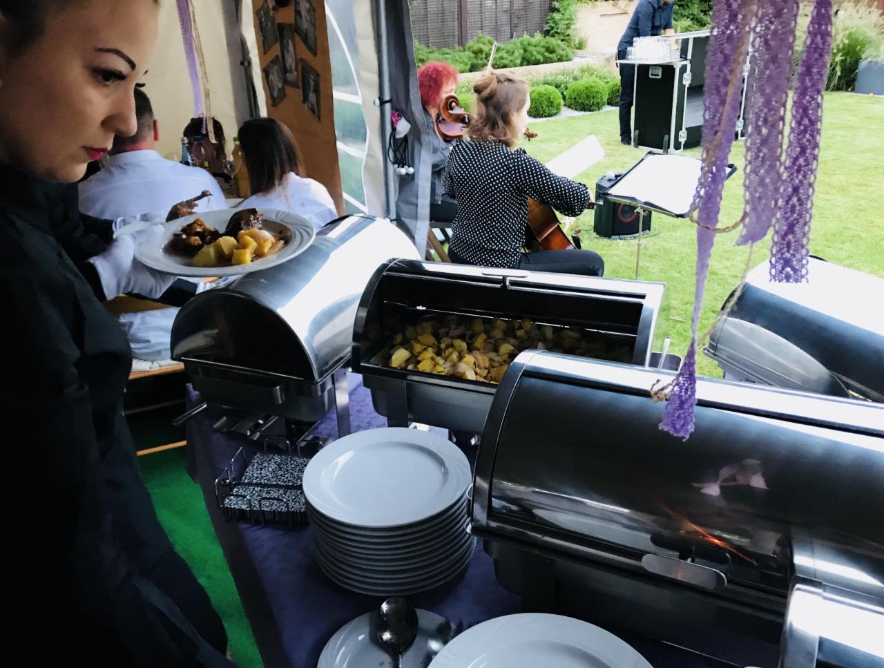 Catering na zewnątrz z muzyką na żywo: bufet z podgrzewaczami, stosy talerzy i kelnerka podająca dania, w tle skrzypaczka i perkusista.