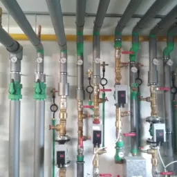 Złożony system rur grzewczych i hydraulicznych z zaworami, manometrami i sterownikami, widoczne rury szare, zielone i miedziane, instalacja przy ścianie.