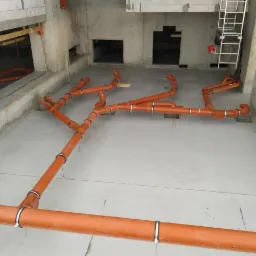 Pomarańczowe rury kanalizacyjne zamontowane na surowym betonowym podłożu wewnątrz budynku w trakcie budowy, widoczna drabina i otwory w ścianach.
