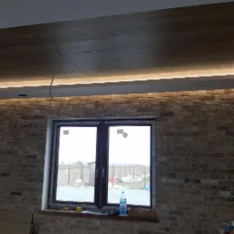 Nowoczesne oświetlenie LED w suficie podwieszanym z drewnopodobną okleiną, ściana z cegły dekoracyjnej, okno z widokiem na zewnątrz, wnętrze w trakcie wykończenia.