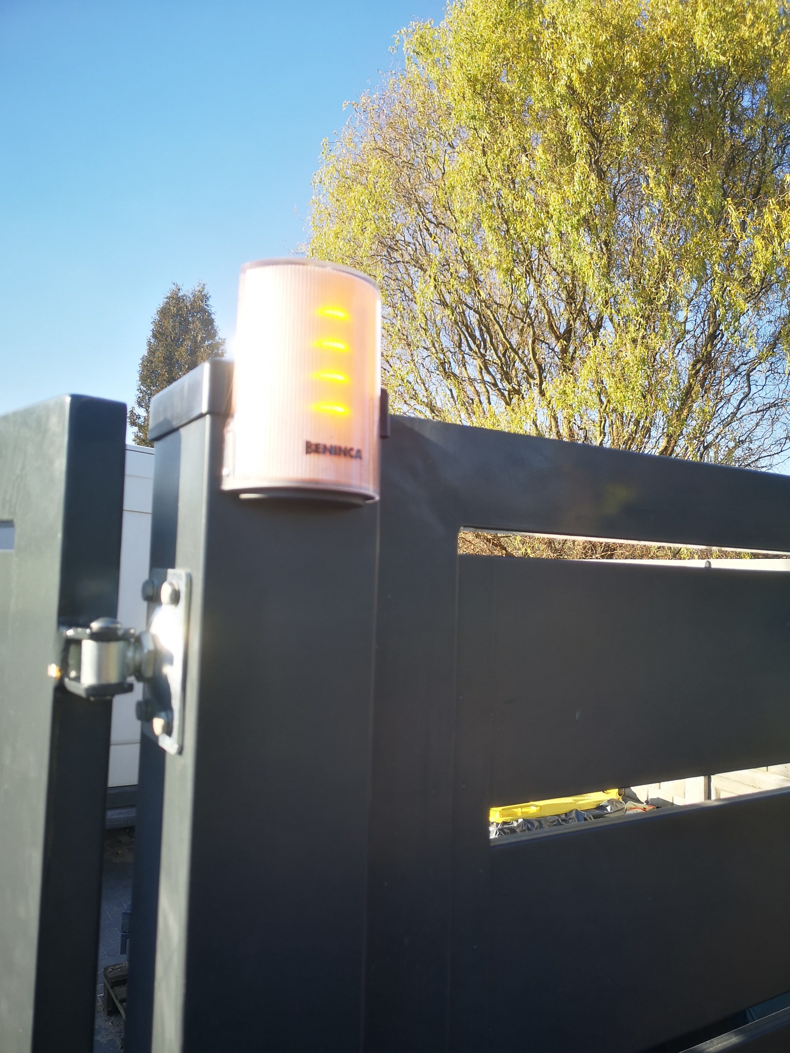 Lampa ostrzegawcza Beninca na czarnej, nowoczesnej bramie wjazdowej, z zielonym drzewem w tle i błękitnym niebem. Widoczne detale montażu.