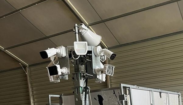 Kompleksowy system monitoringu wizyjnego z kamerami, reflektorami i głośnikiem na metalowej konstrukcji, umieszczony wewnątrz hali z blachy falistej.