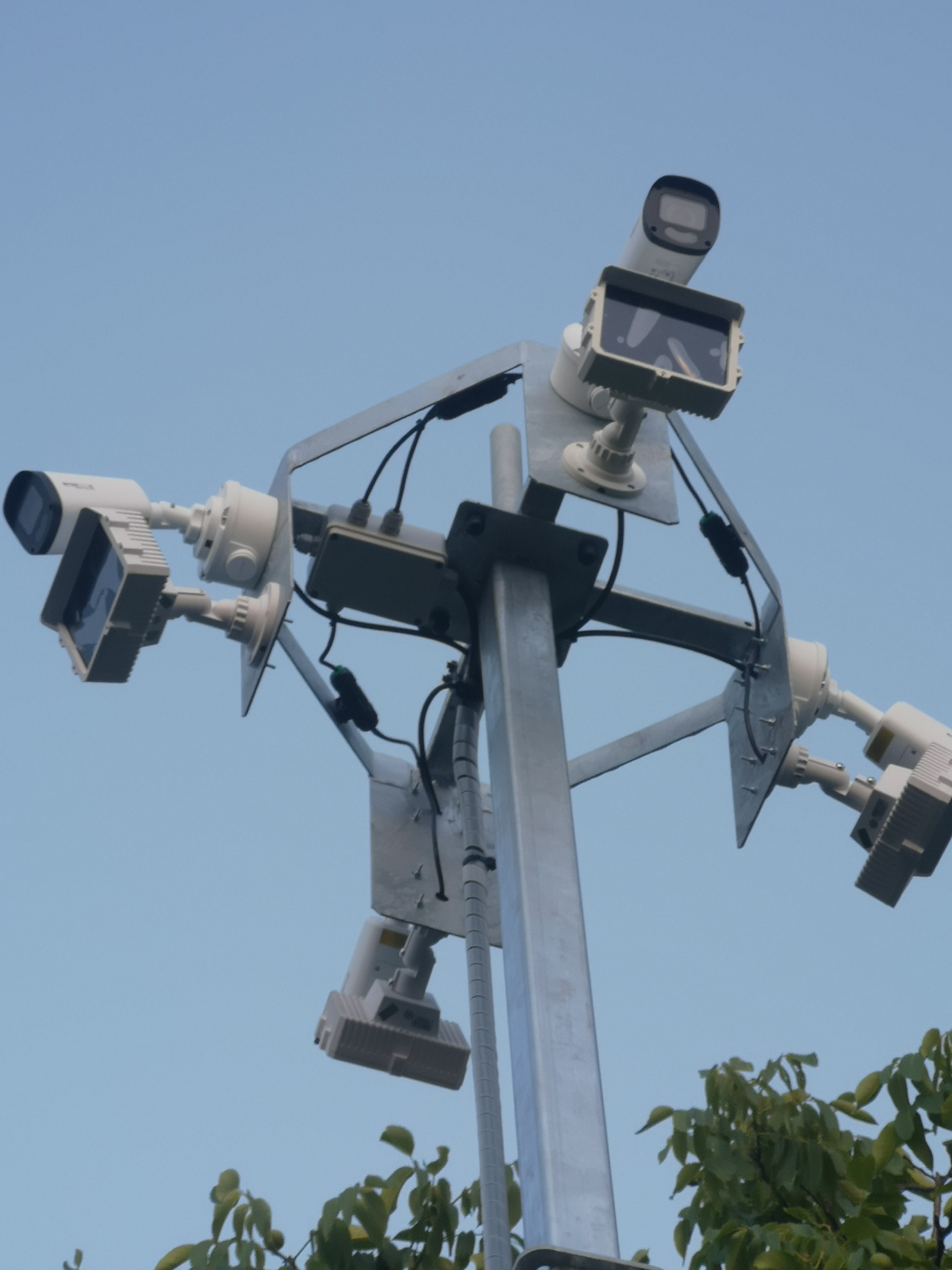 Słup z zamontowanymi kamerami monitoringu skierowanymi w różne strony, widok z dołu na tle błękitnego nieba i fragmentu zieleni.