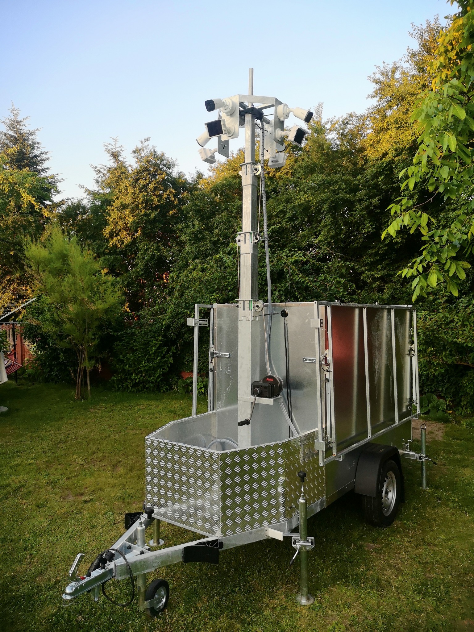 Mobilna wieża monitoringu na przyczepie z wieloma kamerami, ustawiona na trawniku, otoczona zielenią. Widoczne elementy konstrukcyjne i zabezpieczenia.